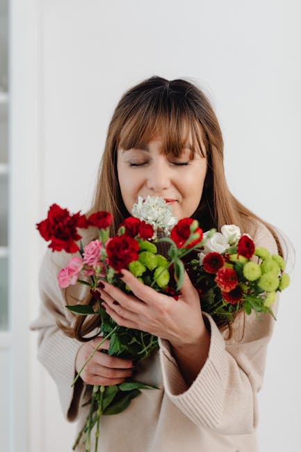 A woman with long brown hair and a light beige sweater is holding a vibrant bouquet of mixed flowers, including red carnations, pink roses, white hydrangeas, and green chrysanthemums, arranged with lu