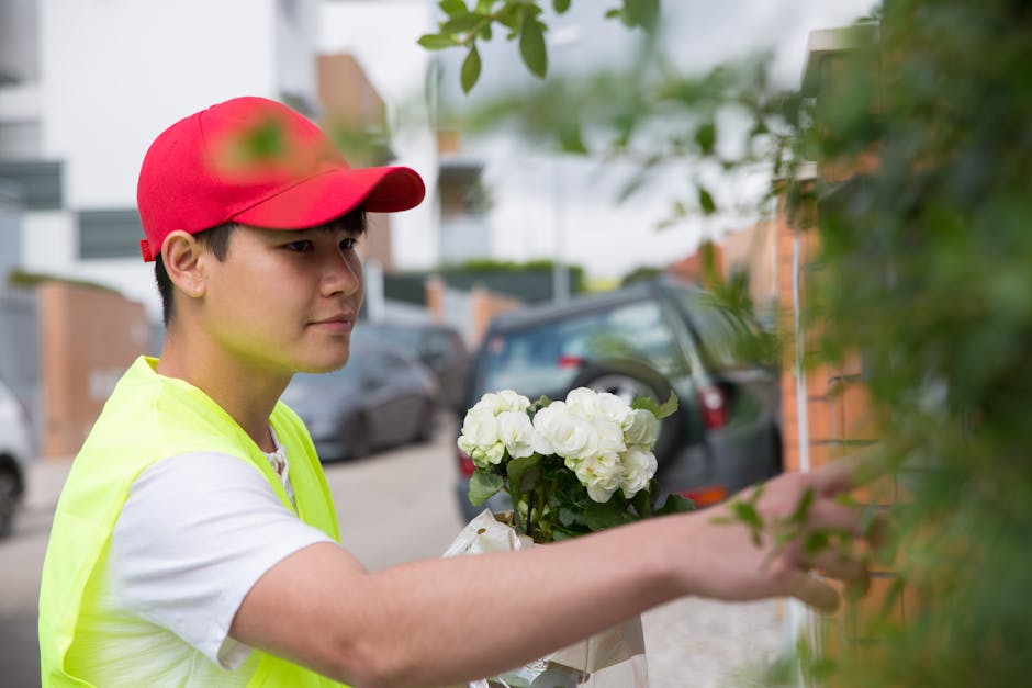 A young male flower delivery person wearing a red cap and a fluorescent yellow vest is placing a bouquet of fresh white roses and hydrangeas against a shrub outside a residential area. The bouquet is 