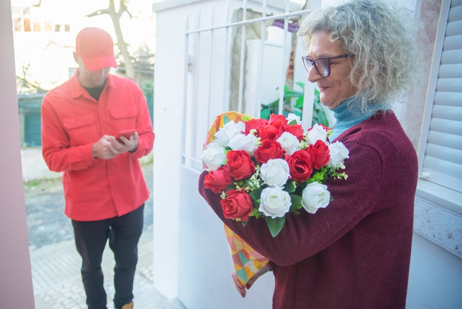 A woman with curly gray hair, wearing glasses, a maroon sweater, and a light blue turtleneck, is holding a vibrant bouquet of fresh red and white roses wrapped in colorful paper. She is smiling gently