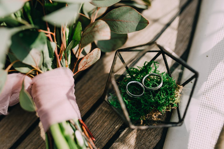 A close-up of a bouquet with greenery and eucalyptus leaves, wrapped with a soft pink ribbon, resting on a wooden surface. Next to it is a geometric glass terrarium containing bright green moss and tw