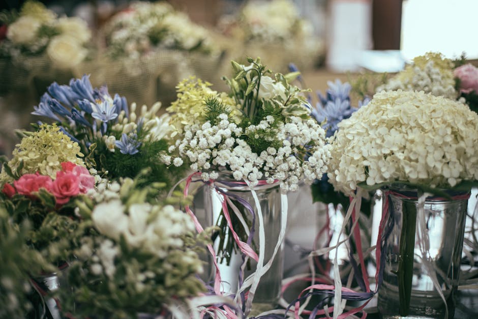 A collection of floral arrangements featuring white hydrangeas, pink roses, and blue and white delphiniums displayed in clear glass vases decorated with pink and white ribbons. The flowers appear fres